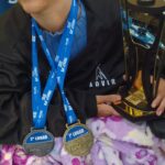 Licélia está sorrindo, usando touca cinza e jaqueta preta com o logo da ADVIR (Associação de Deficientes Visuais de Rio do Sul). Ela segura um troféu dourado com asas estilizadas e exibe no pescoço duas medalhas: uma de 1º lugar e outra de 2º lugar, conquistadas na Copa SESC Santa Catarina. Ela está sentada, coberta com uma manta lilás florida.