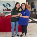 Lucineide está de camisa azul escura, recebendo seu troféu dourado ao lado de uma mulher de cabelo escuro e camisa verde da organização do torneio. Ambas sorriem. Atrás delas, há uma faixa com o logo da ACACE (Associação Caruaruense de Cegos). A premiação ocorreu no 1º Torneio Rápido de Xadrez para Deficientes Visuais de Caruaru.