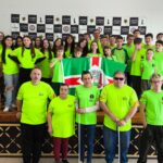 Grupo de aproximadamente 40 pessoas posando para foto em um palco durante um evento de xadrez. Todos vestem camisetas verde-limão com logotipos da Federação de Xadrez e outras marcas. Em destaque, cinco pessoas estão à frente do palco, incluindo duas com bengalas brancas, indicando deficiência visual. Ao centro, duas crianças seguram uma bandeira com o brasão de uma cidade, predominante nas cores verde, branca e vermelha. Ao fundo, um banner com logotipos relacionados ao xadrez. Ambiente iluminado e com piso xadrez em parte do palco.