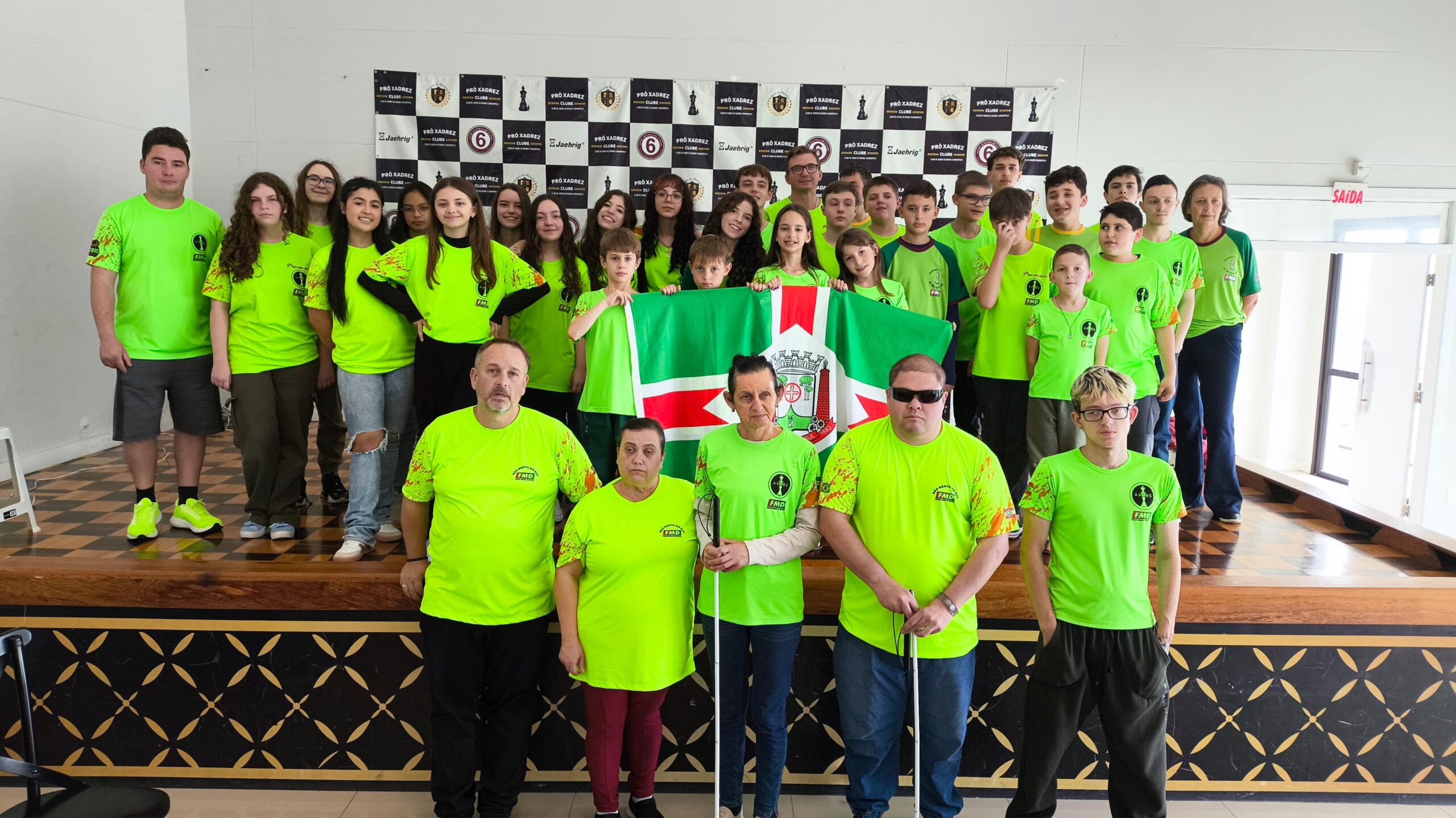 Grupo de aproximadamente 40 pessoas posando para foto em um palco durante um evento de xadrez. Todos vestem camisetas verde-limão com logotipos da Federação de Xadrez e outras marcas. Em destaque, cinco pessoas estão à frente do palco, incluindo duas com bengalas brancas, indicando deficiência visual. Ao centro, duas crianças seguram uma bandeira com o brasão de uma cidade, predominante nas cores verde, branca e vermelha. Ao fundo, um banner com logotipos relacionados ao xadrez. Ambiente iluminado e com piso xadrez em parte do palco.