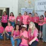 Um grupo de cerca de 20 mulheres em ambiente interno. Algumas estão em pé e outras sentadas ou agachadas no chão. Elas usam camiseta rosa com desenho de uma coroa preta acima das letras “MXDV” e abaixo, está escrito “Mulheres EnXadristas com Deficiência Visual”. Todas estão com medalhas em fita rosa penduradas no pescoço e algumas seguram troféus. Ao fundo, em uma parede de pedra, há uma faixa grande com o texto "Torneio Feminino de Xadrez para Mulheres com Deficiência Visual" e um cartaz do "2º Torneio Presencial da MXDV do Brasil".