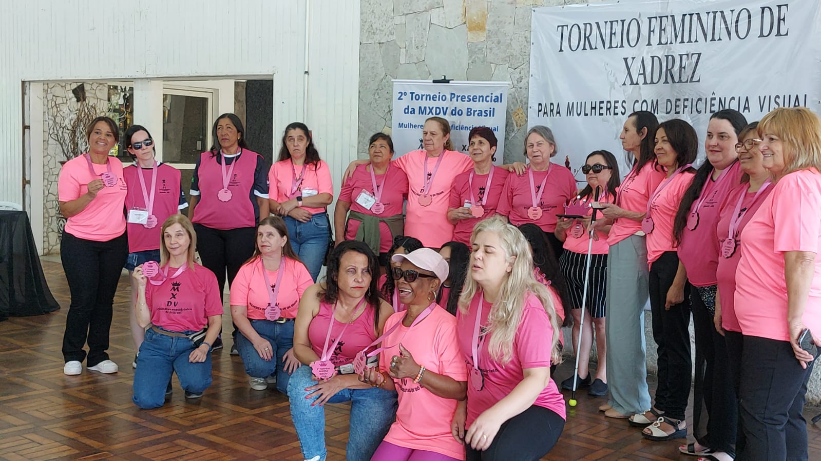 Um grupo de cerca de 20 mulheres em ambiente interno. Algumas estão em pé e outras sentadas ou agachadas no chão. Elas usam camiseta rosa com desenho de uma coroa preta acima das letras “MXDV” e abaixo, está escrito “Mulheres EnXadristas com Deficiência Visual”. Todas estão com medalhas em fita rosa penduradas no pescoço e algumas seguram troféus. Ao fundo, em uma parede de pedra, há uma faixa grande com o texto "Torneio Feminino de Xadrez para Mulheres com Deficiência Visual" e um cartaz do "2º Torneio Presencial da MXDV do Brasil".