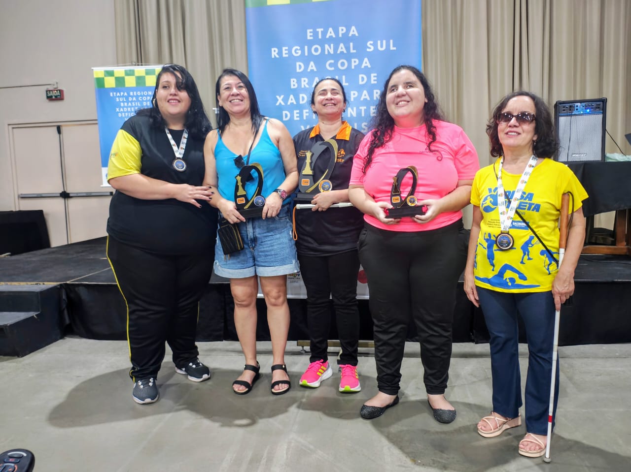 Jaciara, Marcia, Glenda, Brenda e Glaucia estão em pé sorrindo, segurando troféus e medalhas, em frente a um banner escrito Etapa Regional Sul da Copa Brasil de xadrez para deficientes visuais em clima de conquista e celebração. Classificação feminina 1 - Glenda Felippe e Silva – 4 pontos 2 - Márcia Maria Dias Lopes – 4 pontos 3 - Brenda de Paula Pires – 3,5 pontos 4 - Jaciara Marciane da Rosa – 3,5 pontos 5 - Gláucia Teixeira de Queiroz – 3 pontos