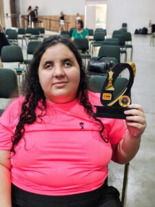 Brenda está sentada no salão de jogos, vestindo uma camiseta rosa vibrante com o símbolo da MXDV. Ela segura com firmeza um troféu preto com detalhes dourado de 3º lugar com símbolo de xadrez, demonstrando conquista e orgulho. Seu olhar transmite tranquilidade e satisfação, como quem reconhece o esforço que valeu a pena. Ao fundo, o ambiente com cadeiras e algumas pessoas reforça o clima de evento, enquanto o destaque permanece nela e em sua vitória.”