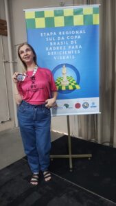 Eni Kindermann é uma mulher loira com os cabelos soltos, lisos na altura dos ombros, em pé, camiseta de mangas curtas rosa com um símbolo de coroa e o texto MXDV “ Mulheres enxadristas Dv do Brasil”, calça jeans azul, sorrindo e segurando uma medalha próxima ao peito. Ao fundo, um banner com detalhes quadriculados acima nas cores verde e amarelo e ao meio escrito Etapa Regional Sul da Copa Brasil de Xadrez para deficientes visuais.