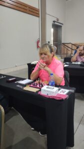 Gercilia está sentada à mesa em uma partida de xadrez em andamento, vestindo uma camiseta rosa da MXDV “Mulheres Enxadristas com Deficiência Visual do Brasil. Com as mãos entrelaçadas e o olhar fixo para frente, ela demonstra total concentração, como se cada movimento estivesse sendo cuidadosamente pensado. À sua frente, as peças estão organizadas sob o tabuleiro. Sobre a mesa, há um relógio de xadrez, uma garrafa de água e uma caixa com o nome ‘Gercilia’, indicando sua identificação no evento. Ao fundo, outras pessoas também participam, reforçando o clima silencioso e tenso de competição. A cena transmite foco, estratégia e a força de quem está ali dando o seu melhor.”