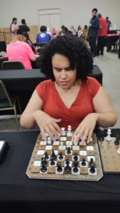 Lais está sentada diante de um tabuleiro de xadrez. Ela tem cabelos curtos, cacheados e escuros, e veste uma blusa vermelha. Sua expressão é concentrada e séria, com o olhar voltado para o tabuleiro. Suas mãos estão posicionadas sobre as peças, explorando cuidadosamente cada posição com delicadeza e precisão. O ambiente com várias pessoas ao fundo conversam, observam ou também participam do torneio, criando um ambiente movimentado, mas organizado. A cena transmite foco, estratégia e inclusão, destacando a habilidade e a dedicação da jogadora no momento da partida. Há algo forte e bonito nesse momento: não é apenas uma partida de xadrez, é superação, habilidade e paixão pelo xadrez.