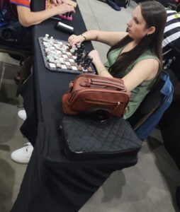 Em meio ao silêncio concentrado de uma partida, Lara está sentada diante de seu tabuleiro de xadrez. Seus cabelos longos e lisos caem sobre os ombros, e ela veste uma blusa verde. Seu olhar está fixo para frente, profundo, como se enxergasse muito além do que está ali. Com a mão delicadamente suspensa, ela toca uma peça, decidindo o próximo movimento. É um gesto simples, mas carregado de tensão, estratégia e sentimento. O tempo parece desacelerar naquele instante. Sobre a mesa, duas bolsas (Uma preta do tabuleiro e uma marrom de uso pessoal) repousam, testemunhas silenciosas daquela disputa intensa. Cada jogada ali não é só um movimento — é raciocínio, é coragem, é a busca por superação. A cena transmite foco e emoção, mostrando que, no xadrez, assim como na vida, cada escolha tem um peso… e cada decisão pode mudar tudo.