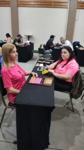 Eni (á esquerda a) e Brenda (á direita) estão sentadas frente a frente em uma mesa em uma partida xadrez da etapa Regional Sul. Ambas usam camisetas rosa com identificação MXDV, elas carregam algo em comum… muito além do uniforme. Eni de cabelos claros e lisos, com postura ereta e expressão tranquila, enquanto Brenda, de cabelos escuros e ondulados, está concentrada, com a mão próxima a um relógio de xadrez amarelo e o tabuleiro entre elas é adaptado, com peças encaixadas, e há também uma caixa de peças ao lado. As duas estão focadas na partida, não como um confronto qualquer…mas como um encontro de trajetórias… de histórias… de dedicação, cada movimento é pensado… sentido… respeitado. Não há rivalidade e sim admiração e companheirismo… mesmo em lados opostos do tabuleiro. Ao fundo, outras pessoas também estão sentadas em mesas, participando de jogos ou, em um ambiente organizado e silencioso.