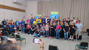 Um grande grupo de pessoas está reunido, posando para uma foto. Elas estão organizadas em várias fileiras, algumas em pé, outras sentadas ou agachadas à frente. É um grupo de pessoas com deficiência visual utilizando bengalas e também uma pessoa em cadeira de rodas. Muitos vestem camisetas de equipes, nas cores azul, preto e laranja, Rosa pink e alguns usam medalhas no pescoço. No centro, algumas pessoas seguram um troféu e uma bandeira amarela com o símbolo da sua equipe. Ao fundo, há banners com referência a Etapa Regional Sul da Copa Brasil de Xadrez para deficientes visuais. Na frente da cena, duas pessoas seguram celulares, registrando o momento. O ambiente é um salão amplo, com cadeiras espalhadas e uma cortina grande ao fundo. A cena transmite um momento coletivo de celebração e conquista.